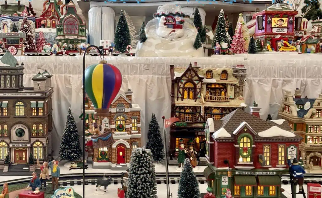 A festive miniature holiday village display featuring detailed buildings, a hot air balloon, snowy trees, and figurines on a train set.